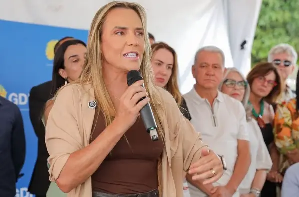Celina Leão falando ao microfone cercada de pessoas em um evento