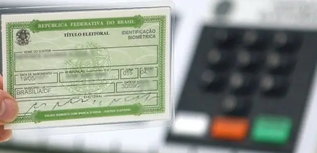 Mão segurando um título de eleitor diante de uma urna eletrônica