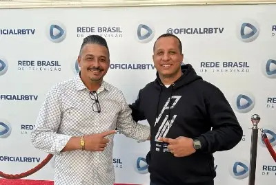 Dois homens posando diante de painel da Rede Brasil de Televisão