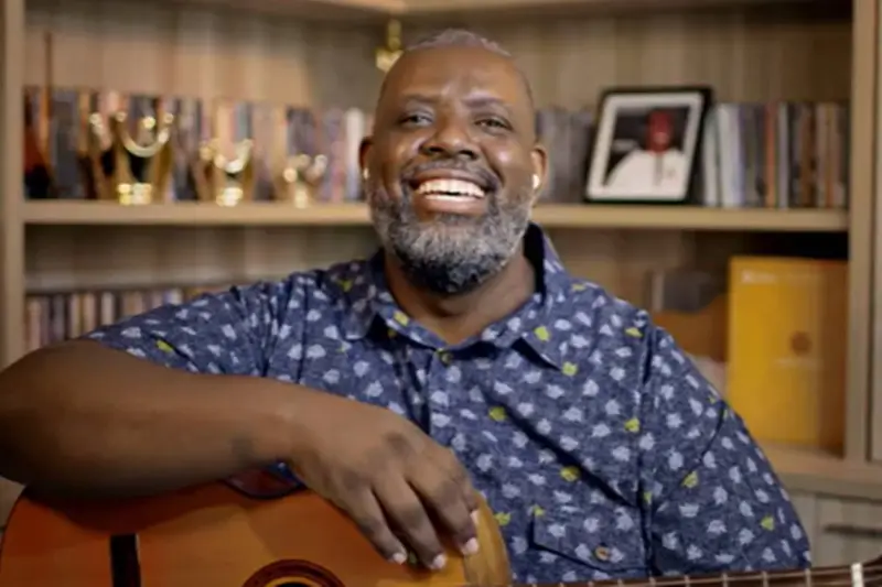 Músico sorrindo com um violão diante de uma estante